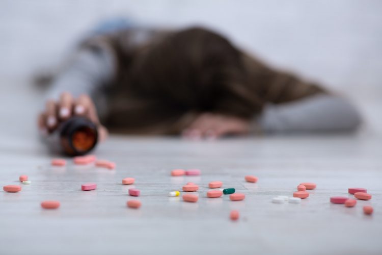 Young woman committing suicide, lying on floor with tablets scattered from jar, selective focus