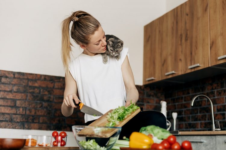 Young woman preparing vegetarian green salad at home and playing with cat