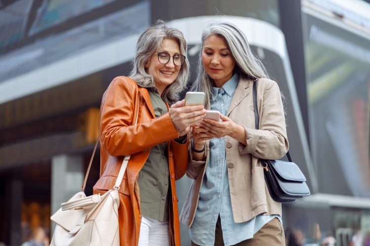 Positive grey haired ladies look at mobile phones on modern city street