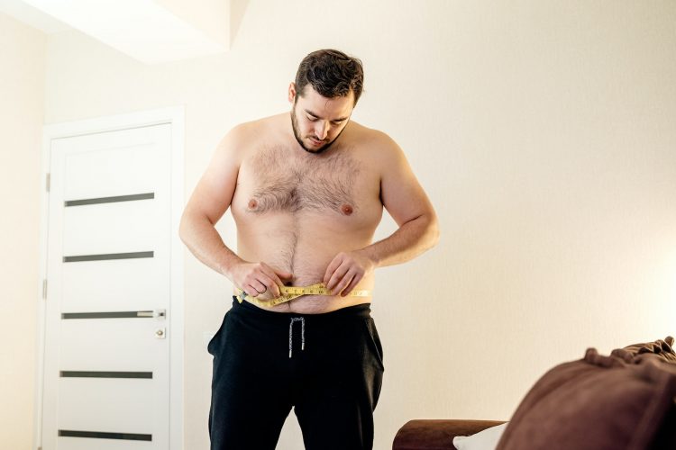 Fat sad male measuring his belly with measurement tape standing in the living room