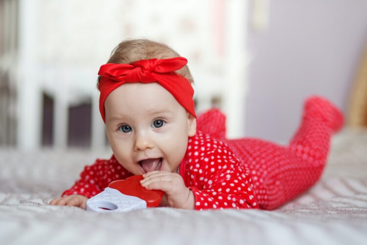 Little baby 6 months old in red sleepsuit and headband. The baby is played with rattle.