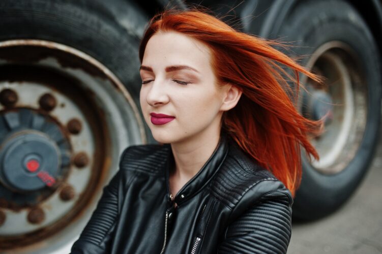Red haired stylish girl wear in black, sitting against large truck wheels.