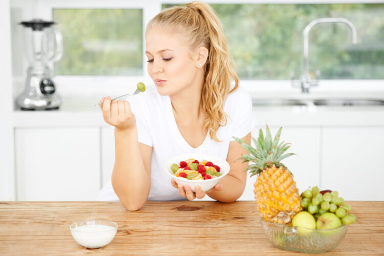 Mmmm, tasty. Attractive young woman eating fruit salad in her kitchen.