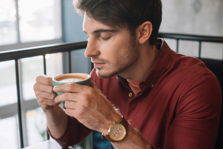 young man smelling cappuccino with closed eyes on balcony in coffee shop